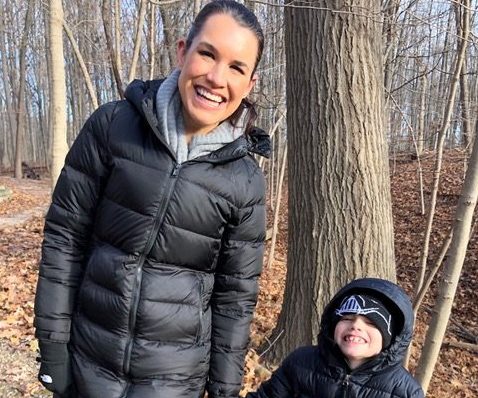 Tall woman with dark hair dressed in winter jacket walks hand in hand with a small boy, her son