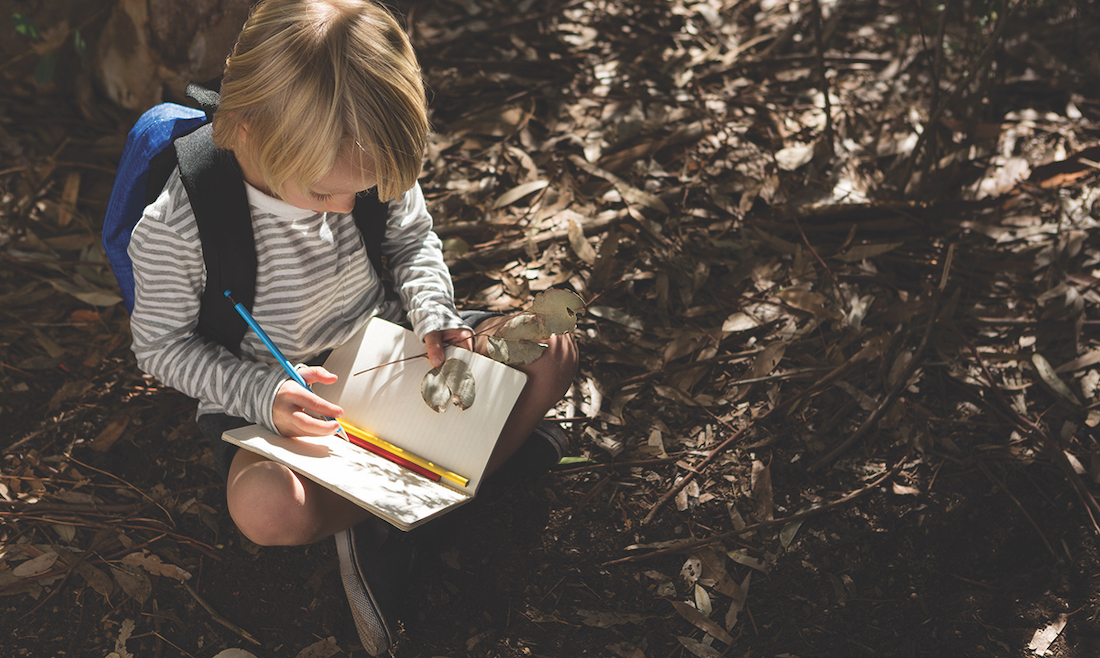 Little girl with backpack sitting on ground writing in notebook